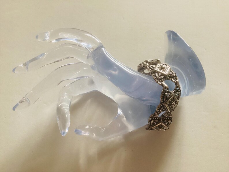 May include: Silver bracelet with a geometric design. The bracelet is made of multiple linked pieces with a square shape in the center of each piece.