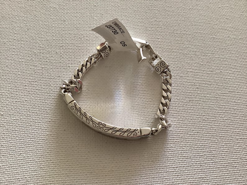 K&ouml;nnte beinhalten: Ein silbernes Armband mit einem Panzerketten-Design. Das Armband hat einen dekorativen Stab mit einem eingravierten Blattmuster. Ein Etikett ist am Armband befestigt mit dem Wort "EMBRACE" und der Nummer "J37730".