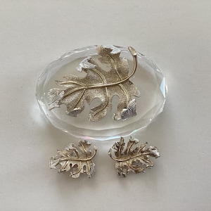 Puede incluir: Un broche de metal plateado y un par de pendientes de clip a juego, cada uno con forma de hoja de roble texturizada con una vena central prominente. El broche se muestra descansando sobre un pisapapeles de cristal transparente, ovalado y facetado.