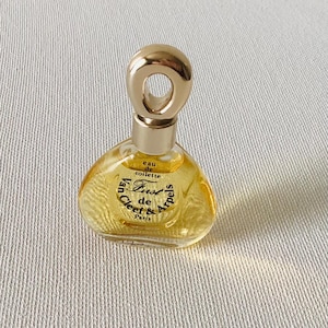 May include: A miniature glass perfume bottle containing a golden liquid, featuring a rounded triangular shape and a gold-toned cap with a circular opening. The label reads: eau de toilette, Van Cleef & Arpels, First de, Paris.