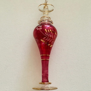 Puede incluir: Frasco de perfume de vidrio rojo rubí con detalles dorados. El frasco tiene un cuerpo bulboso con diseños florales grabados y un tallo delgado. La parte superior presenta un tapón de vidrio transparente con borde dorado y un pequeño lazo.