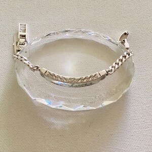 K&ouml;nnte beinhalten: Ein silbernes Armband mit Kettendesign und einem gebogenen Steg, auf dem das Wort "embrace" und Bl&auml;ttermuster eingraviert sind. Das Armband wird auf einem klaren, facettierten Kristall pr&auml;sentiert. Schmuck.
