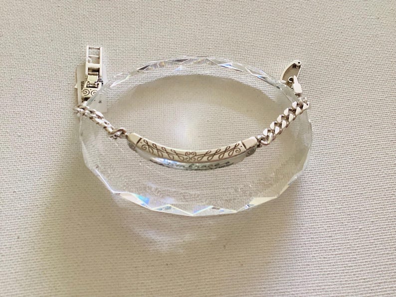 K&ouml;nnte beinhalten: Ein silbernes Armband mit Kettendesign und einem gebogenen Steg, auf dem das Wort "embrace" und Bl&auml;ttermuster eingraviert sind. Das Armband wird auf einem klaren, facettierten Kristall pr&auml;sentiert. Schmuck.