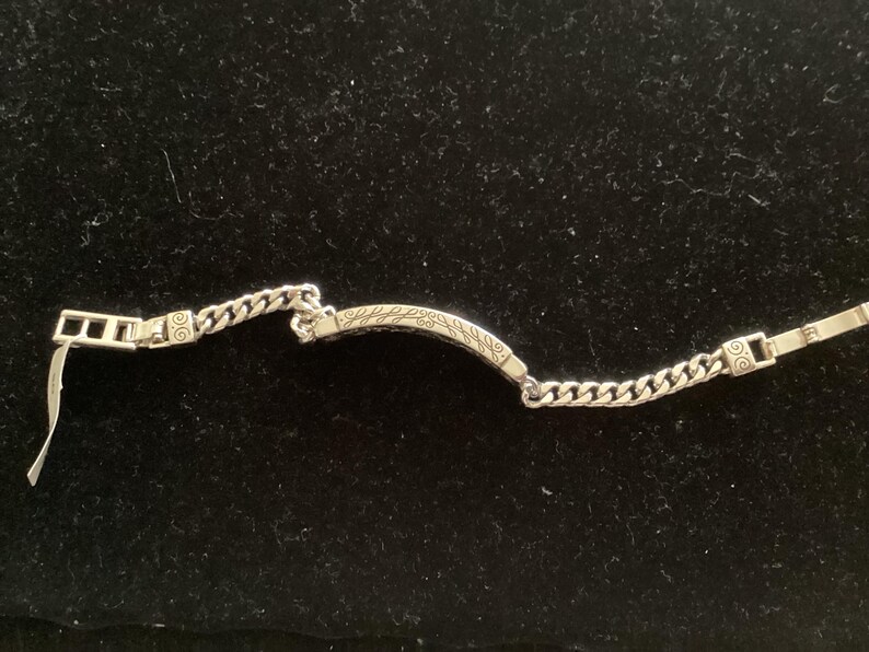 K&ouml;nnte beinhalten: Silberfarbenes Armband mit einer Panzerkette und einem dekorativen, gebogenen Steg. Der Steg hat ein eingraviertes Blattmuster. Das Armband hat einen rechteckigen Verschluss und liegt auf schwarzem Untergrund.