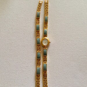 May include: A gold-toned wristwatch with a white face and a matching bracelet. The bracelet features rectangular, light green stones and decorative gold links. The watch and bracelet are displayed on a neutral, textured surface.