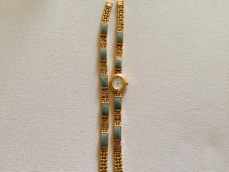 May include: A gold-toned wristwatch with a white face and a matching bracelet. The bracelet features rectangular, light green stones and decorative gold links. The watch and bracelet are displayed on a neutral, textured surface.