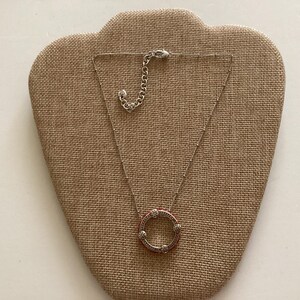 May include: A silver necklace with a round pendant featuring a circle of small white stones and a red center. The necklace is on a brown fabric display.