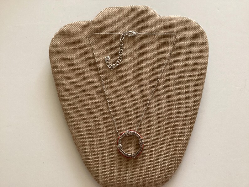 May include: A silver necklace with a round pendant featuring a circle of small white stones and a red center. The necklace is on a brown fabric display.