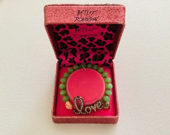 Może przedstawiać: Bransoletka Betsey Johnson z zielonymi fasetowanymi koralikami i złotą zawieszką z napisem Love wysadzaną białymi cyrkoniami. Bransoletka znajduje się w różowym, brokatowym pudełku z wyściółką w panterkę i nadrukowaną nazwą marki.