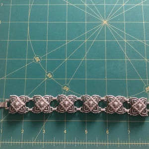 May include: A silver bracelet with a chain link design and square-shaped charms. Each charm has a raised, ornate design and a small, clear stone in the center.