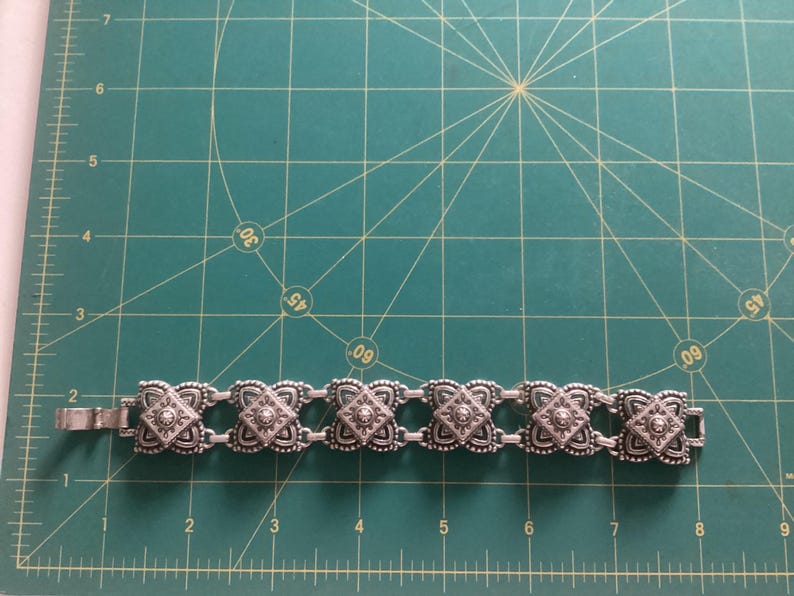 May include: A silver bracelet with a chain link design and square-shaped charms. Each charm has a raised, ornate design and a small, clear stone in the center.