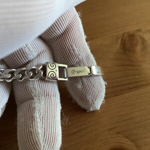 K&ouml;nnte beinhalten: Ein silberfarbenes Armband mit einem Panzerketten-Design. Der Verschluss hat ein dekoratives Wirbelmuster und der Markenname "Brighton" ist eingraviert. Das Armband wird vor einem neutralen Hintergrund gezeigt.