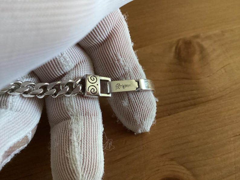 K&ouml;nnte beinhalten: Ein silberfarbenes Armband mit einem Panzerketten-Design. Der Verschluss hat ein dekoratives Wirbelmuster und der Markenname "Brighton" ist eingraviert. Das Armband wird vor einem neutralen Hintergrund gezeigt.