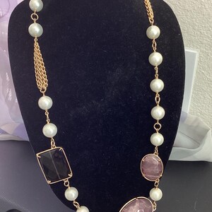 Könnte beinhalten: Eine lange goldfarbene Halskette mit großen Kunstperlen, einem schwarzen quadratischen Stein und zwei ovalen rosa Steinen. Die Halskette hat eine Kette und ein Quastendesign, das auf einer schwarzen Schaufensterpuppe präsentiert wird.