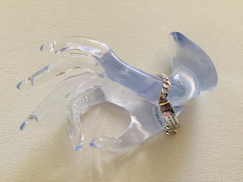 K&ouml;nnte beinhalten: Ein silbernes Armband mit Kettenglied-Design und einem rechteckigen Anh&auml;nger. Der Anh&auml;nger tr&auml;gt die Worte "Forever & Always" und ein Blumenmuster. Das Armband wird auf einem transparenten, handf&ouml;rmigen Display pr&auml;sentiert.