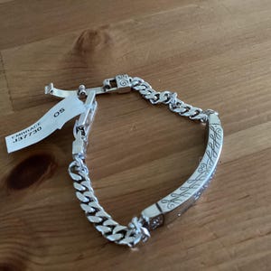 K&ouml;nnte beinhalten: Ein silbernes Armband mit einem Panzerketten-Design und einem gravierten rechteckigen Mittelst&uuml;ck. Das Mittelst&uuml;ck weist ein verziertes Muster auf. Ein Etikett mit dem Text "EMBRACE J37730 OS" ist angebracht.