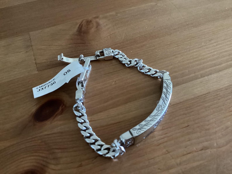 K&ouml;nnte beinhalten: Ein silbernes Armband mit einem Panzerketten-Design und einem gravierten rechteckigen Mittelst&uuml;ck. Das Mittelst&uuml;ck weist ein verziertes Muster auf. Ein Etikett mit dem Text "EMBRACE J37730 OS" ist angebracht.