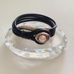 Könnte beinhalten: Ein schwarzes Lederarmband mit silberfarbenem Verschluss und Perlenakzent. Das Armband wird auf einem klaren, facettierten Kristall präsentiert. Das Design ist schlicht und elegant.
