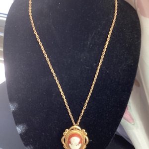 Könnte beinhalten: Eine goldene Kettenkette mit einem Kameenanhänger. Die Kamee ist oval geformt und zeigt ein weißes Profil einer Person auf rotem Hintergrund.