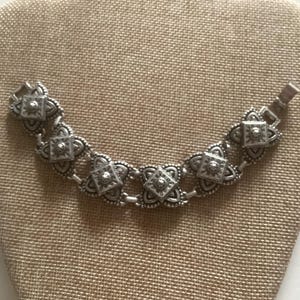 May include: A silver bracelet with a chain link design and ornate, square-shaped charms. The charms have a raised, textured pattern.