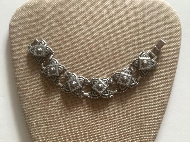 May include: A silver bracelet with a chain link design and ornate, square-shaped charms. The charms have a raised, textured pattern.