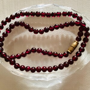 Op de afbeelding: Een ketting van dieprode, ronde kralen met een goudkleurige sluiting, rustend in een heldere, ovaalvormige schaal. De kralen zijn gelijkmatig verdeeld, waardoor een symmetrisch ontwerp ontstaat. De ketting is waarschijnlijk gemaakt van edelstenen.