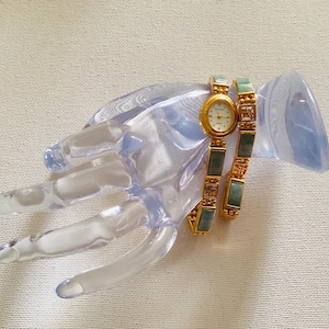 May include: A gold-toned wristwatch with a cream-colored face and a matching bracelet. The bracelet features rectangular green and gold-toned links. The watch and bracelet are displayed on a clear, hand-shaped display stand.