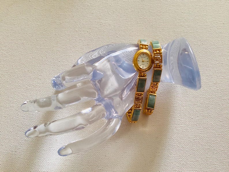 May include: A gold-toned wristwatch with a cream-colored face and a matching bracelet. The bracelet features rectangular green and gold-toned links. The watch and bracelet are displayed on a clear, hand-shaped display stand.