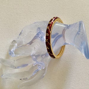 May include: A gold-tone bangle bracelet with a burgundy enamel band and gold floral accents. The bracelet is displayed on a clear, plastic hand-shaped jewelry holder. The background is a light beige fabric.
