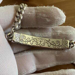 K&ouml;nnte beinhalten: Silberfarbenes Armband mit einer Panzerkette und einem rechteckigen Mittelst&uuml;ck. Das Mittelst&uuml;ck hat ein eingraviertes Blumendesign. Das Armband wird auf einem wei&szlig;en Handschuh gezeigt.