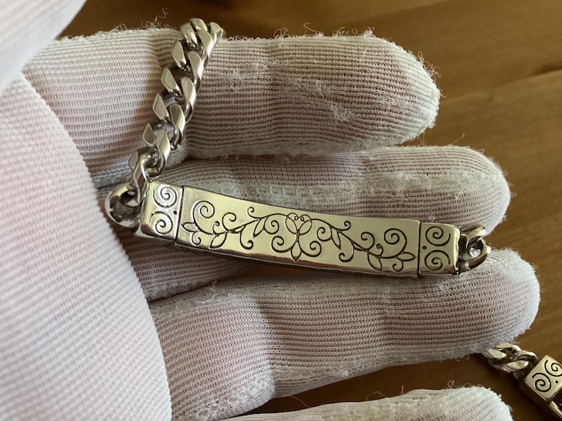 K&ouml;nnte beinhalten: Silberfarbenes Armband mit einer Panzerkette und einem rechteckigen Mittelst&uuml;ck. Das Mittelst&uuml;ck hat ein eingraviertes Blumendesign. Das Armband wird auf einem wei&szlig;en Handschuh gezeigt.