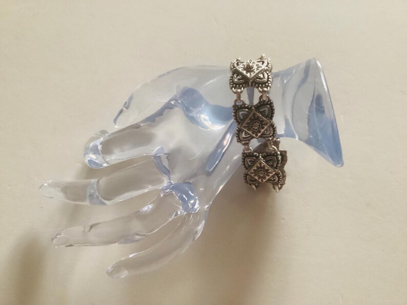 May include: Silver bracelet with a chain link design and intricate square-shaped charms. The charms have a floral pattern and are connected by a delicate chain.