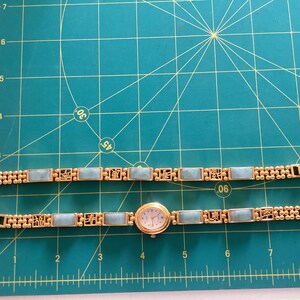 May include: Gold-toned watch and matching bracelet with rectangular light blue stones. The watch has a round face with a white background and gold hands. The bracelet and watch band feature decorative gold filigree.