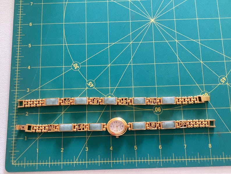 May include: Gold-toned watch and matching bracelet with rectangular light blue stones. The watch has a round face with a white background and gold hands. The bracelet and watch band feature decorative gold filigree.
