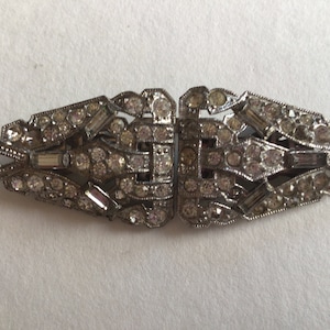 Könnte beinhalten: Eine silberfarbene, geometrische Brosche mit rautenförmigem Design. Die Brosche ist mit klaren Strasssteinen und baguetteförmigen Kristallen verziert.