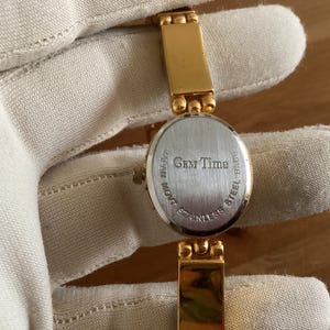 May include: Gold-toned wristwatch with an oval face and a stainless steel back. The watch band is also gold-toned. The watch face has the text "Gem Time" and "Japan Movt".