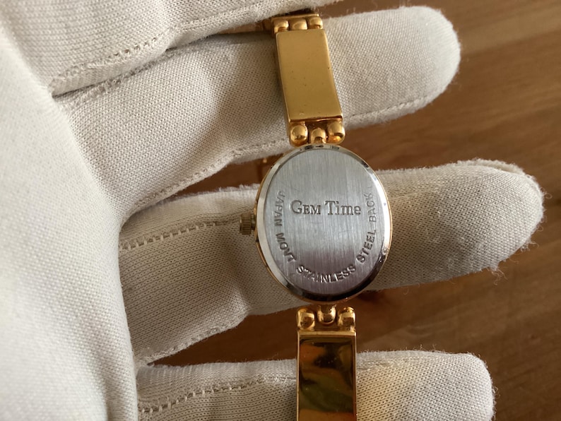 May include: Gold-toned wristwatch with an oval face and a stainless steel back. The watch band is also gold-toned. The watch face has the text "Gem Time" and "Japan Movt".