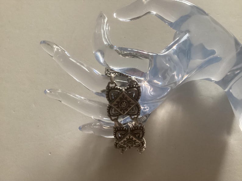 May include: Silver bracelet with a chain link design and decorative square charms. Each charm has a detailed floral pattern.