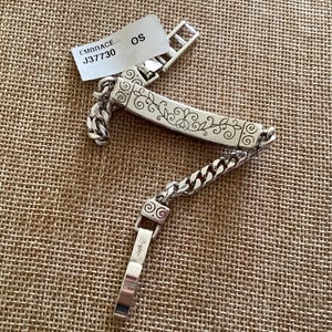 K&ouml;nnte beinhalten: Silberfarbenes Armband mit einem dekorativen floralen Design auf einer rechteckigen Platte. Das Armband hat ein Kettenglied-Design und einen Verschluss. Ein Etikett mit dem Text "EMBRACE... J37730 OS" ist angebracht.