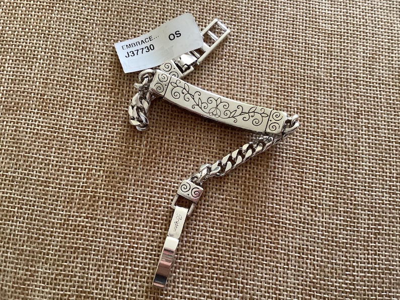 K&ouml;nnte beinhalten: Silberfarbenes Armband mit einem dekorativen floralen Design auf einer rechteckigen Platte. Das Armband hat ein Kettenglied-Design und einen Verschluss. Ein Etikett mit dem Text "EMBRACE... J37730 OS" ist angebracht.