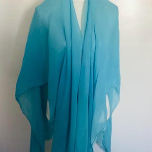 Puede incluir: Un chal azul claro, drapeado sobre un maniquí. La tela tiene una textura fluida y delicada. El diseño presenta mangas anchas y un escote en V profundo, creando una silueta suave y elegante. El chal está hecho de un material ligero.