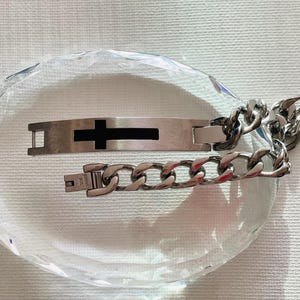 Puede incluir: Una pulsera plateada con un diseño de cruz negra y una cadena de eslabones. La pulsera se muestra sobre una superficie clara y facetada, creando un efecto reflectante. El cierre es visible, y el diseño es moderno y minimalista.