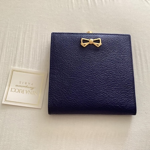 May include: A dark blue square wallet with a textured surface and a gold-toned metal bow emblem on the front. Beside the wallet is a small square white card featuring the text Nina Ricci Paris in gold lettering with a thin gold border.