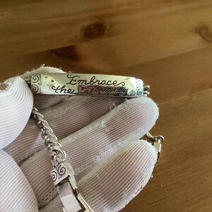 K&ouml;nnte beinhalten: Ein silbernes Armband mit einem rechteckigen Anh&auml;nger, auf dem die Worte "Embrace the Journey" in Schreibschrift eingraviert sind. Das Armband hat ein Kettenglied-Design und wird von einer behandschuhten Hand gehalten.