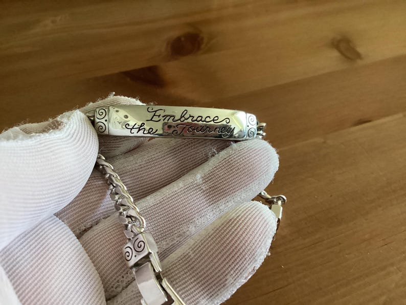 K&ouml;nnte beinhalten: Ein silbernes Armband mit einem rechteckigen Anh&auml;nger, auf dem die Worte "Embrace the Journey" in Schreibschrift eingraviert sind. Das Armband hat ein Kettenglied-Design und wird von einer behandschuhten Hand gehalten.
