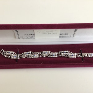 May include: A silver bracelet with blue stones in a red velvet box. The box has a white label that reads "MANUFACTURED It is hereby Certified Mark of and When".