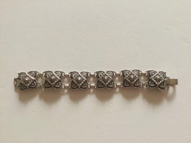 May include: A silver bracelet with a chain link design and square-shaped charms. Each charm has a detailed, intricate pattern.