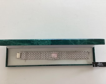 May include: A silver watch with a pink face and a crystal-encrusted band. The watch is displayed in a green velvet box with a white interior.