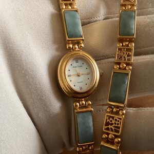 May include: Gold-toned quartz watch with a light blue jade-like stone and a white face. The watch has an oval face with Roman numerals and a matching bracelet. The watch is a quartz movement.