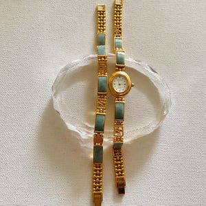 May include: A gold-toned wristwatch with a rectangular jade-colored stone bracelet. The watch face is white with black numerals and hands. The bracelet is made of gold-colored metal with jade-colored rectangular stones. The watch is displayed on a clear, oval-shaped glass.